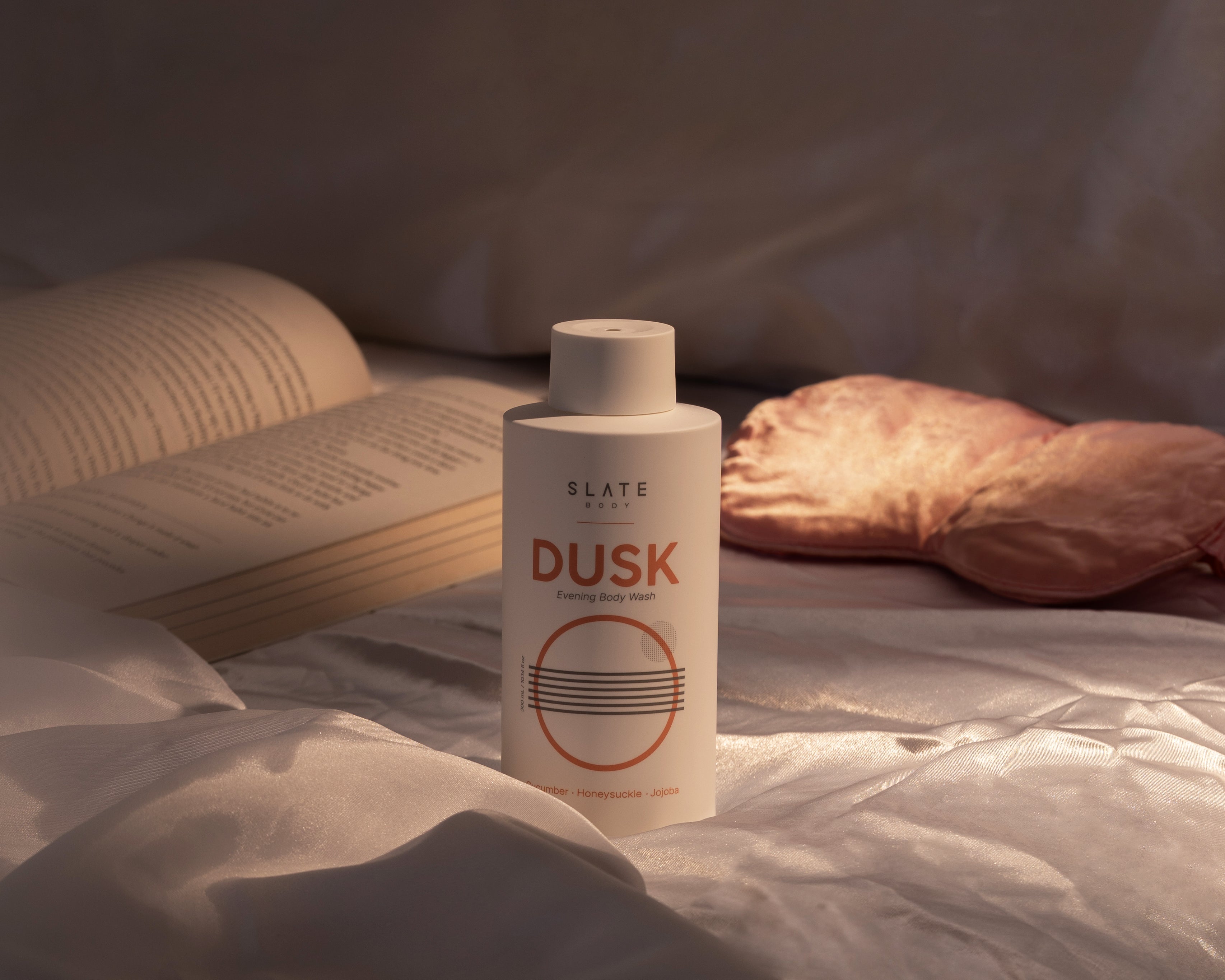 SLARE Body DUSK evening body wash bottle on a bed with an open book and pillow