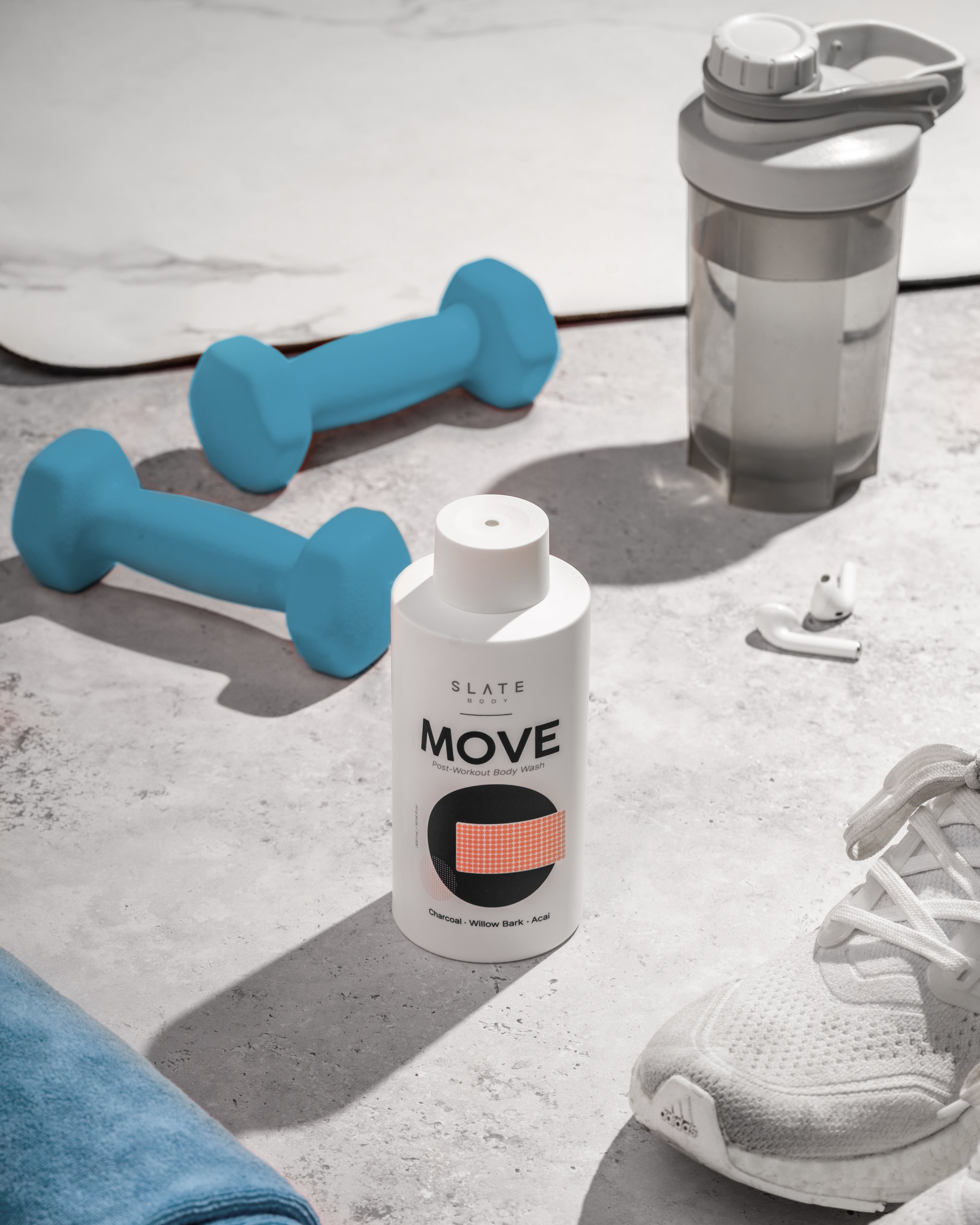 Fitness scene with a bottle of Slate Body MOVE post-workout body wash placed on the floor beside blue dumbbells, a water bottle, and athletic gear