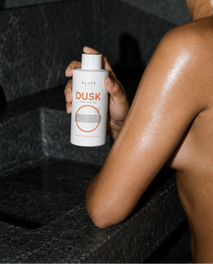 Person holding a bottle labeled 'Dusk' in a bathroom setting - the sexiest body wash for soft skin