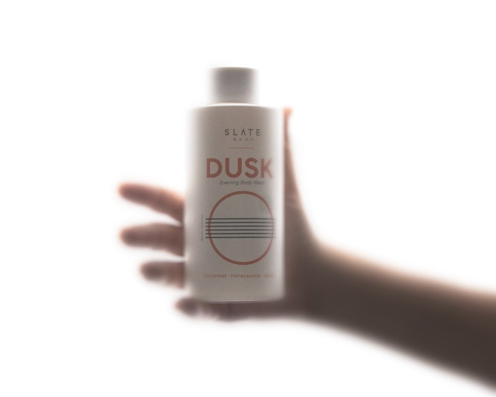A hand holding SLATE Body DUSK Body Wash against a glowing white background. Soft-focus imagery reflects the calming, sensorial mood of an evening shower.