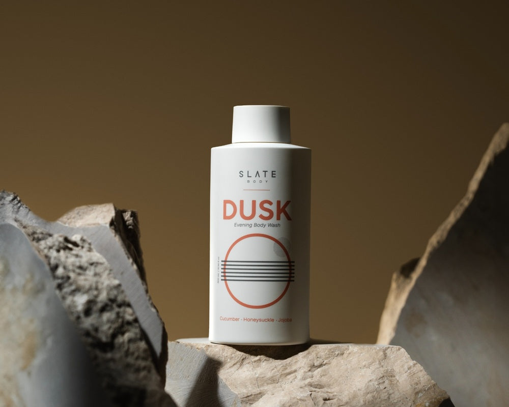 SLATE Body DUSK Body Wash placed on sculpted stones, highlighting the elegant matte bottle with clean orange graphics. Ideal for luxury evening skincare routines.